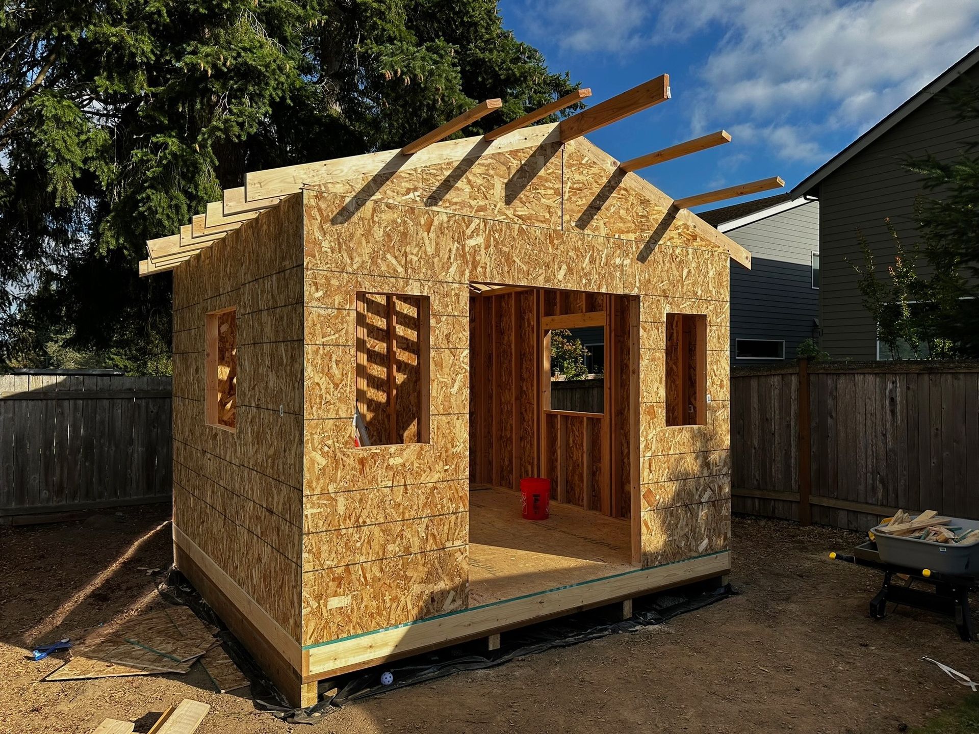 An unfinished wooden shed frame with plywood walls, open door and window frames, standing in a fenced backyard.