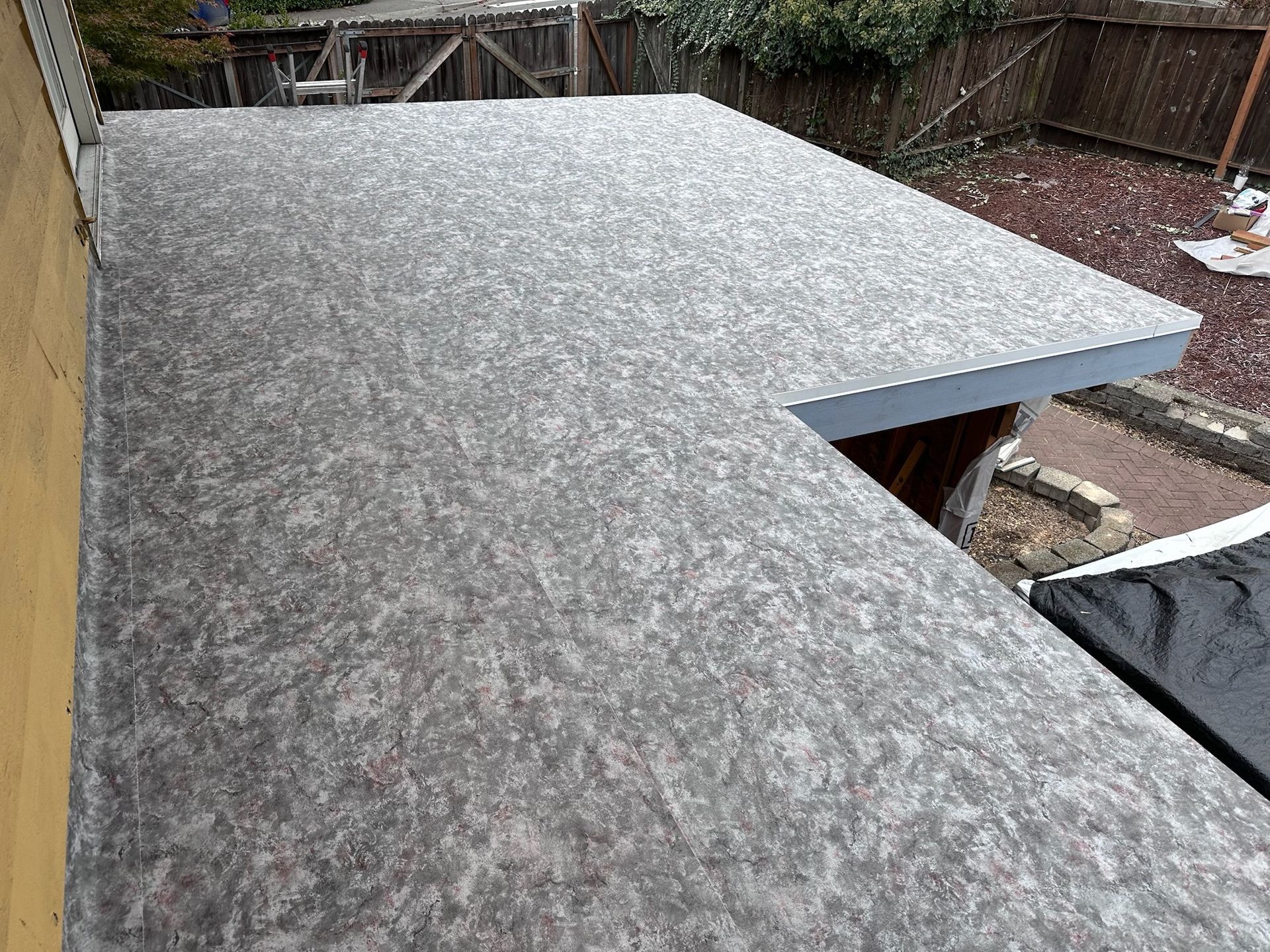 A flat, gray, textured roof surface viewed from above, with a cutout section overlooking a residential backyard.