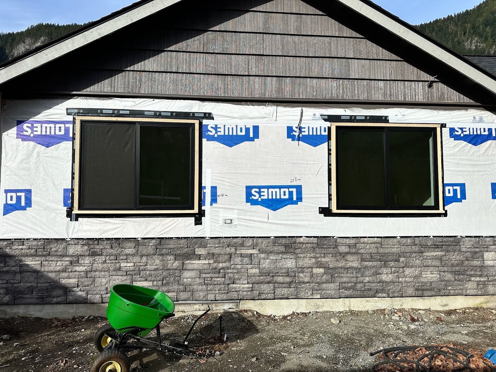 An unfinished house exterior with two installed windows, gray stone siding, and protective wrap, with a green cart below.