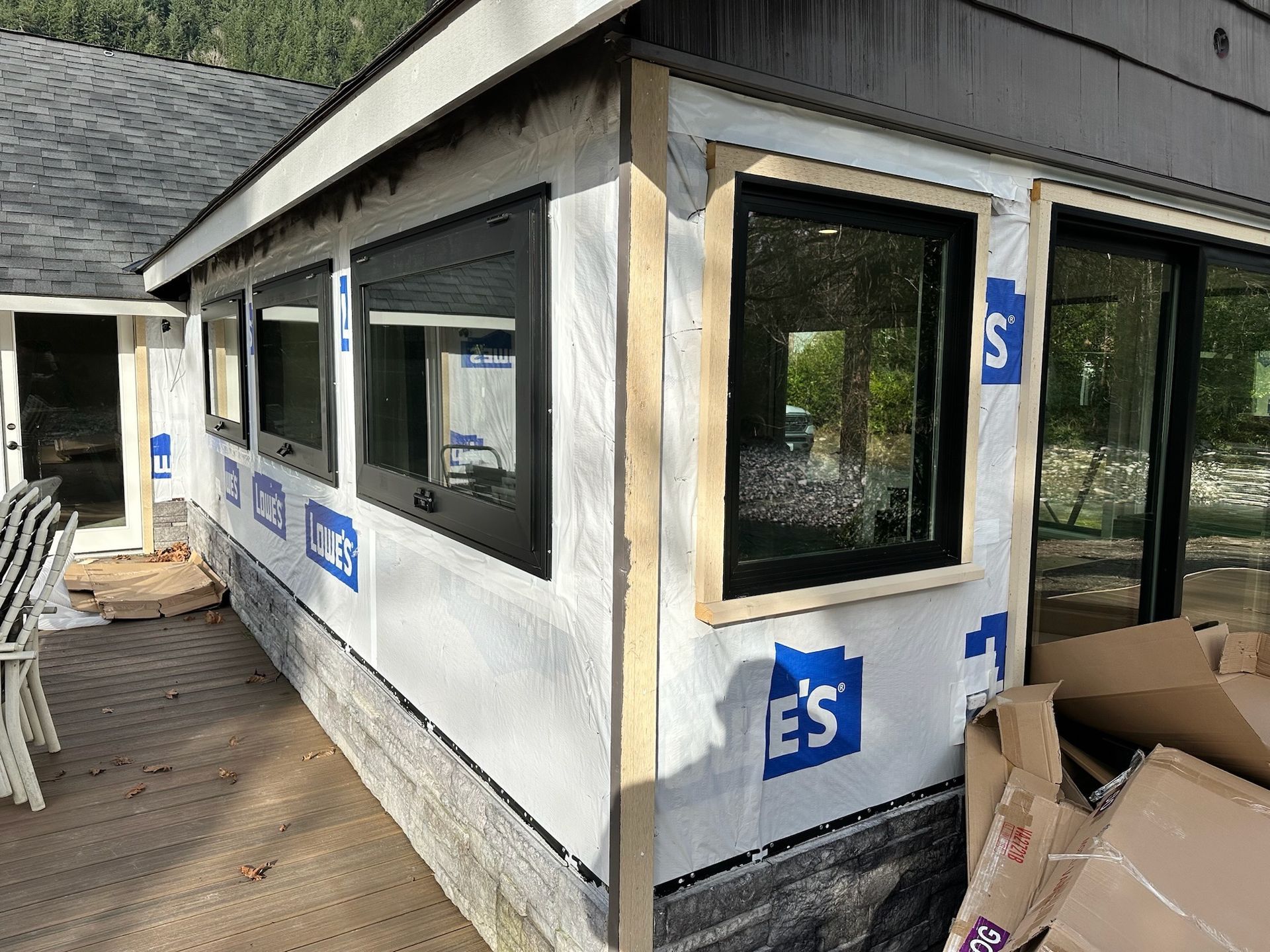 Construction site of a house addition with black-framed windows, white moisture barrier, and a wooden deck.