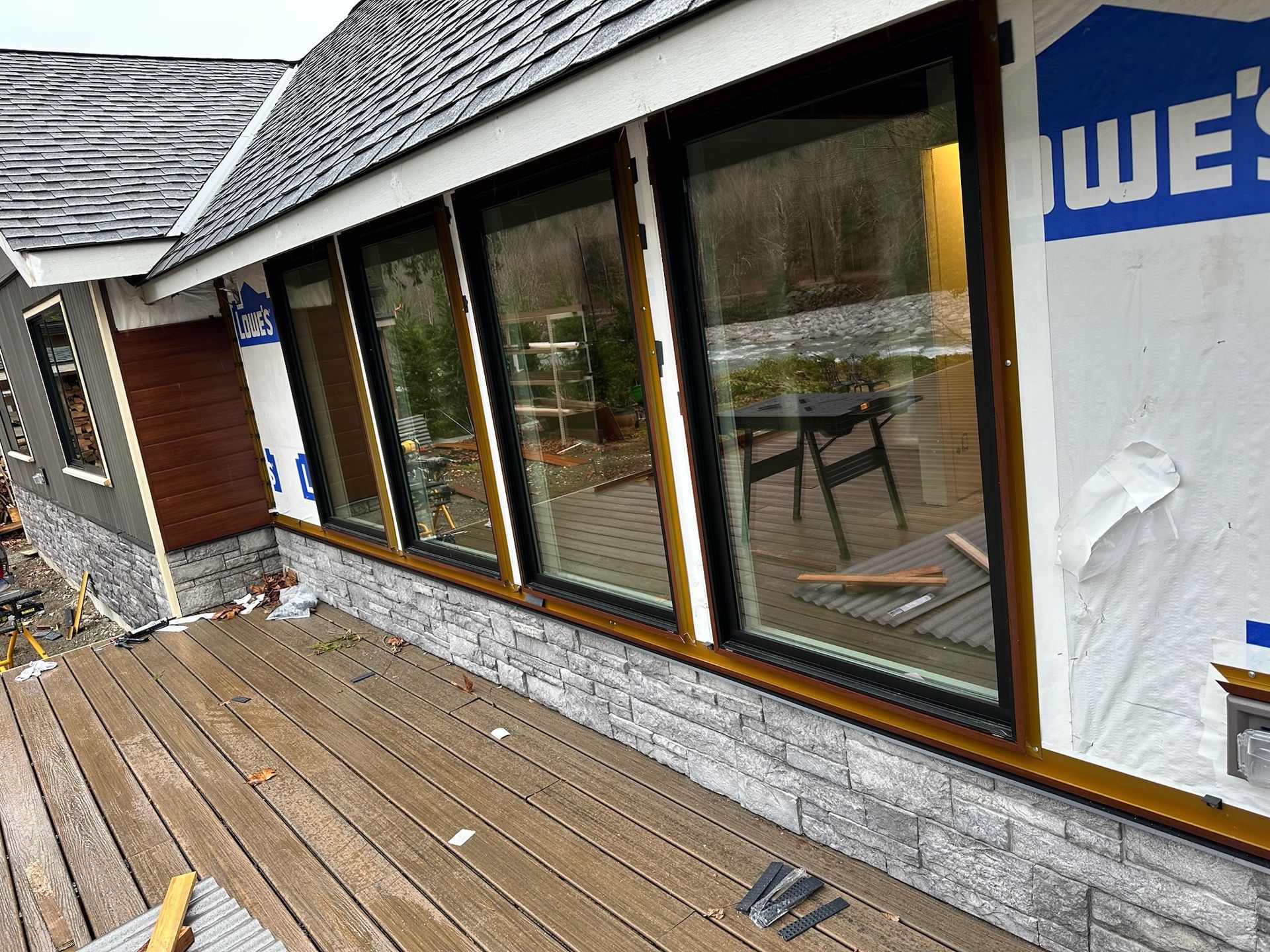Construction site showing a house exterior with a stone foundation, a wooden deck, and large window installations.