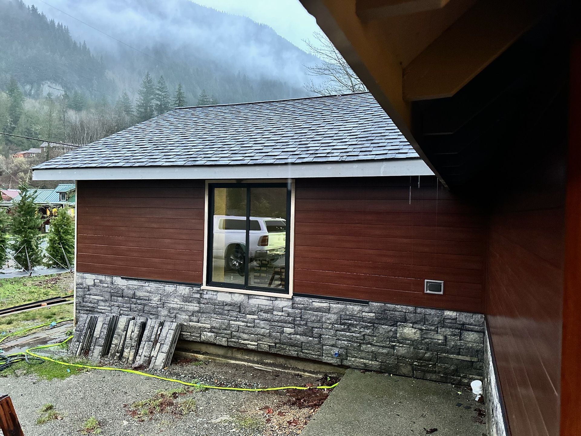 A small, wooden shed with a dark shingled roof and a white window sits in a wooded yard.