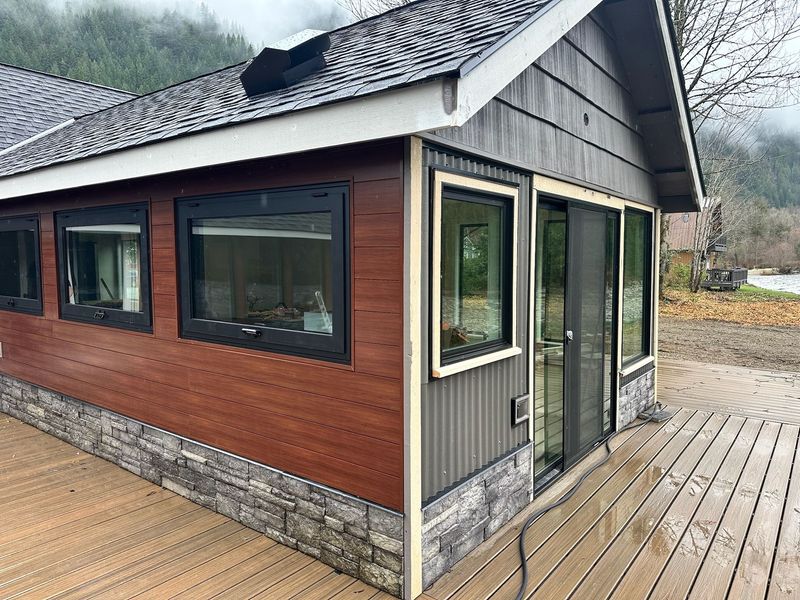 A modern cabin exterior featuring horizontal wood siding, stone base trim, dark-framed windows, and a wooden deck.