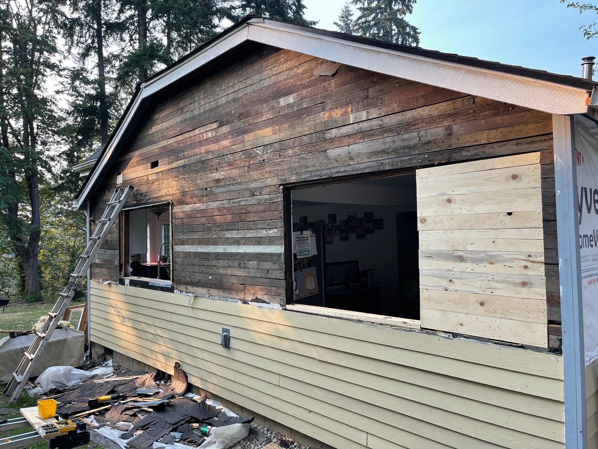 Exterior view of a house during siding installation, showing a mix of old weathered wood, new light siding, and a window.