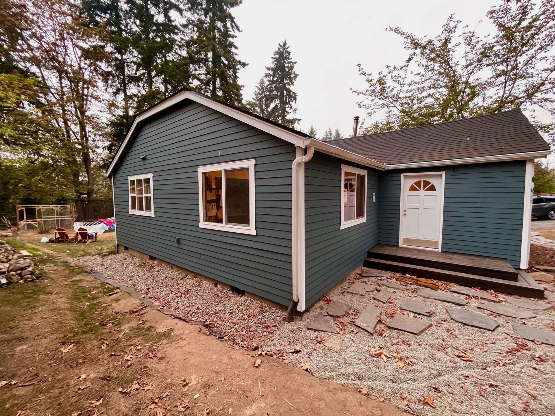 A dark blue, single-story house with white trim, a white front door, and a stone walkway in a wooded, leafy yard.