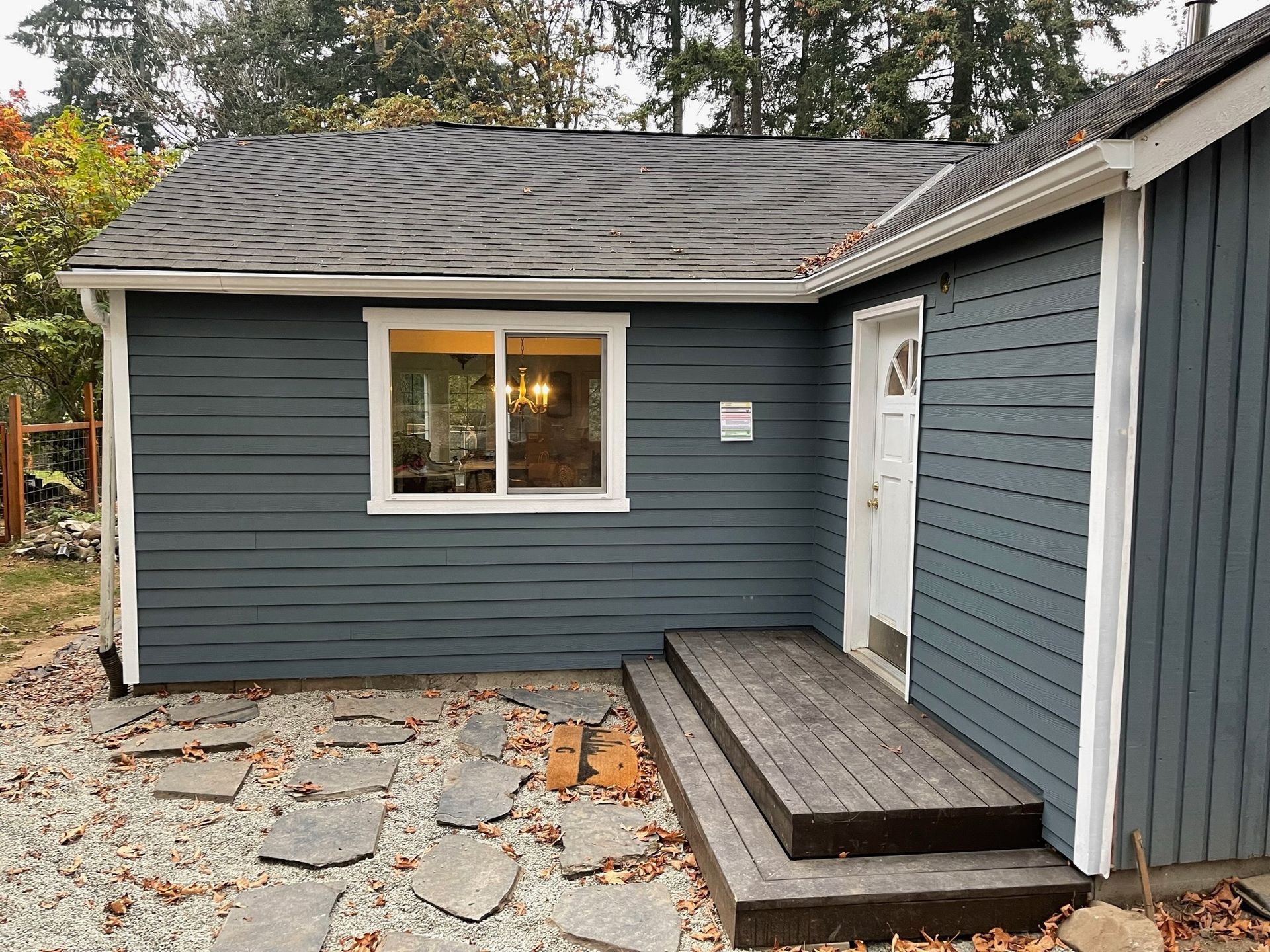 Dark blue house exterior with a white-framed window, a small wooden deck with stairs, and a gravel patio with stone pavers.