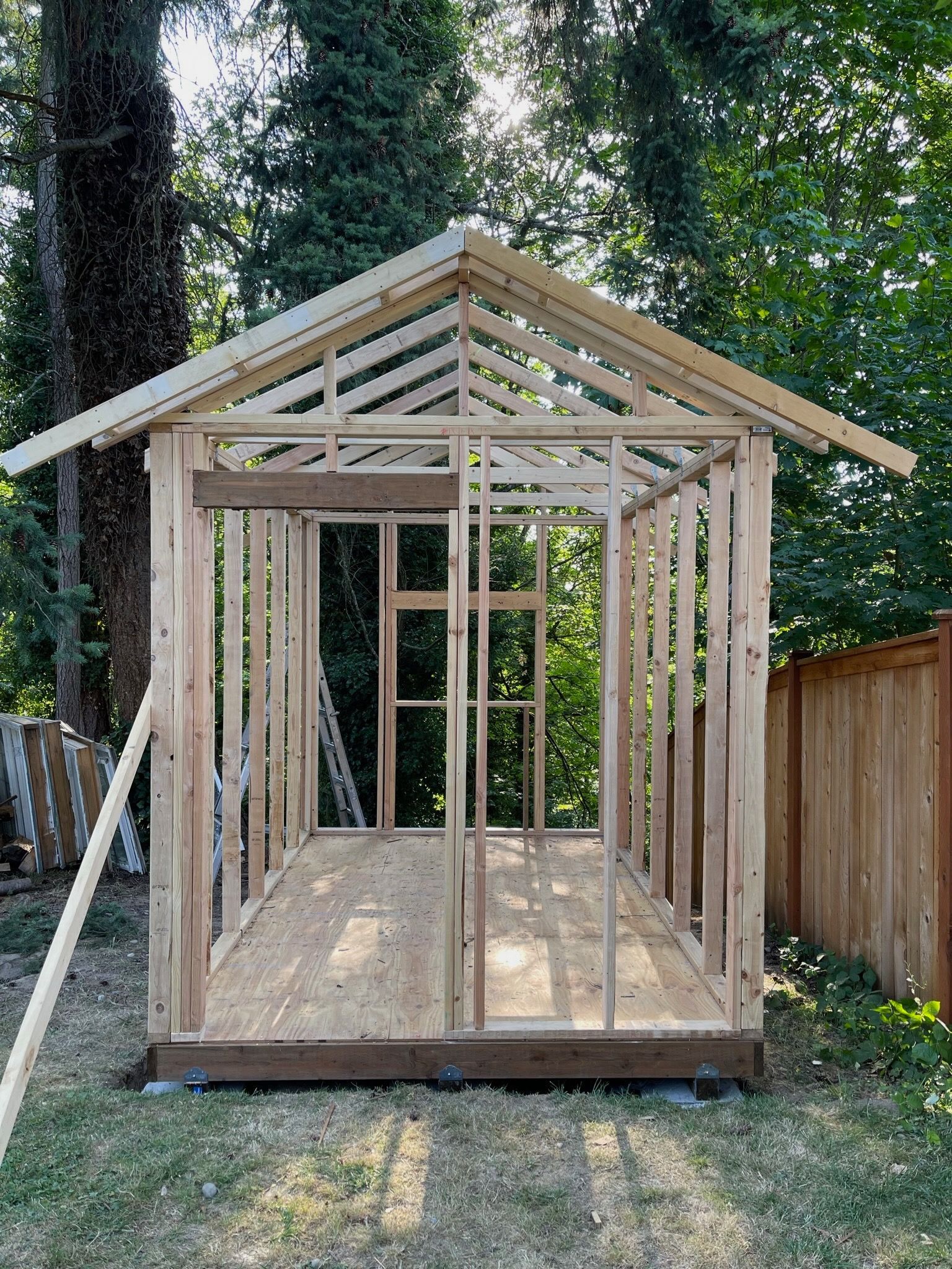 A wooden frame for a small shed under construction, standing on a platform in a yard with trees in the background.