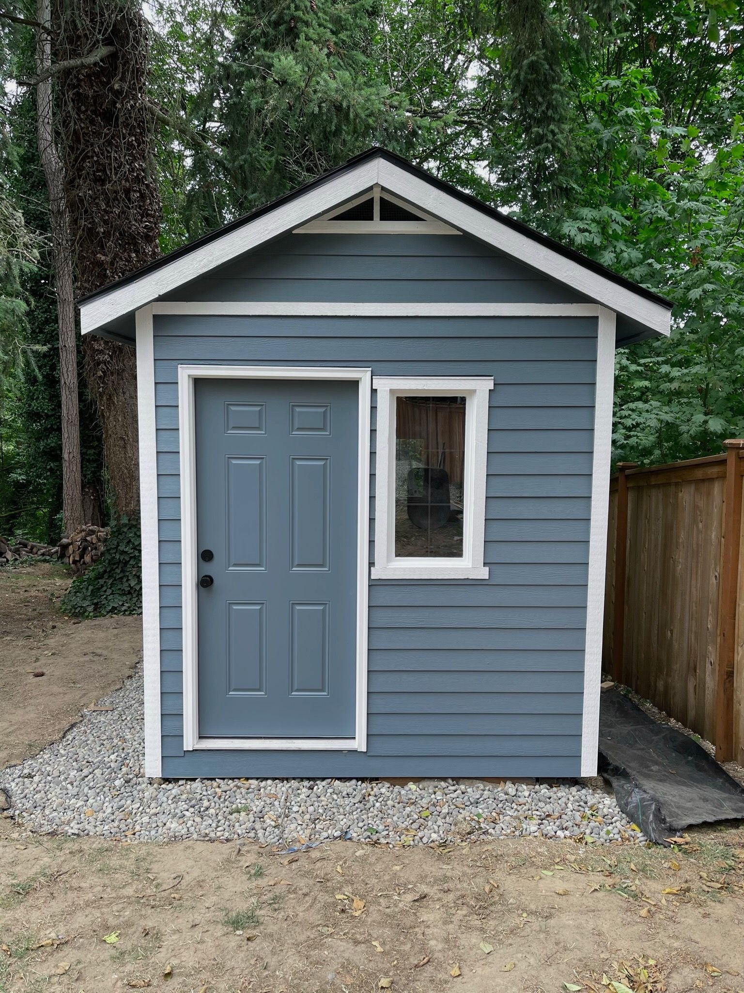 A small, blue-gray wooden shed with a white-trimmed door and window, set on a gravel base in a wooded yard.