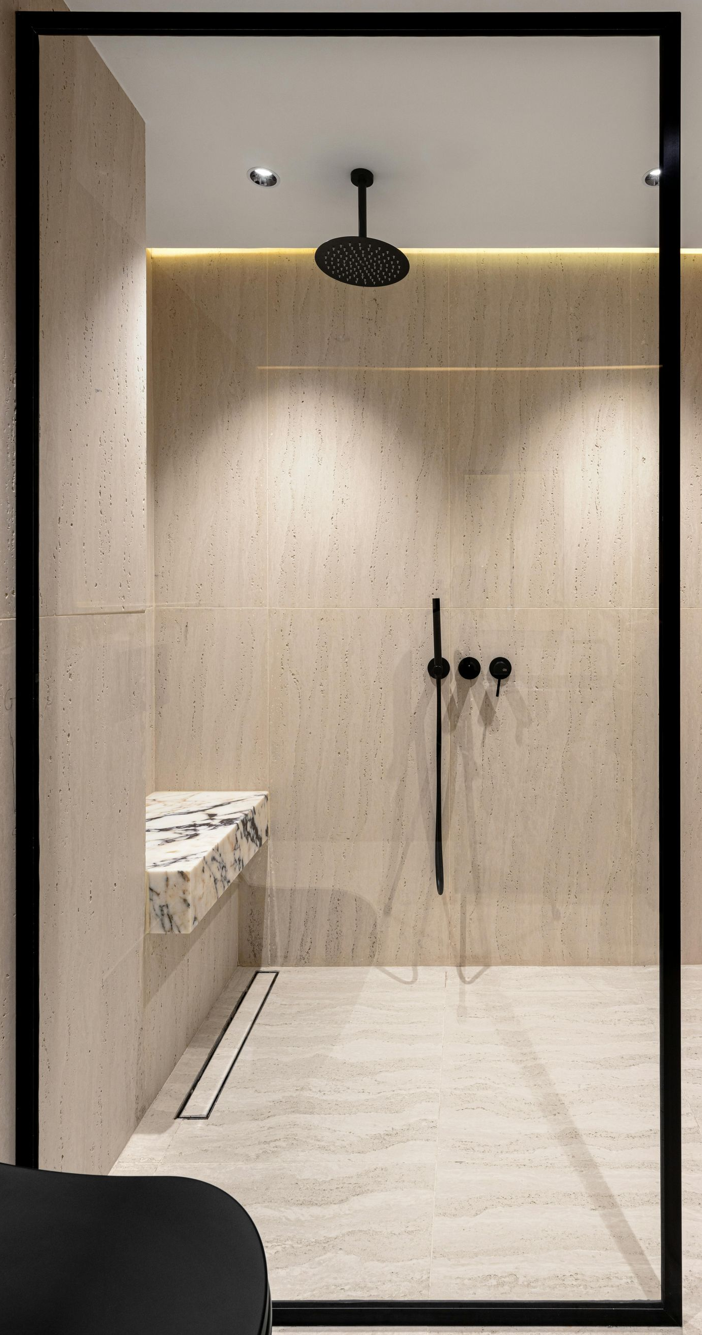 A modern shower with beige stone walls, a black rain showerhead, matching fixtures, and a black-framed glass door.