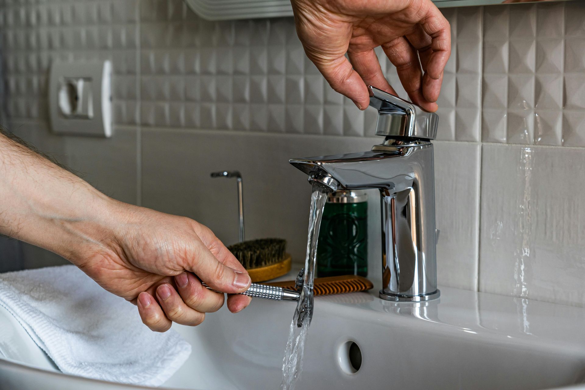 A hand holds a razor under a running chrome bathroom faucet.
