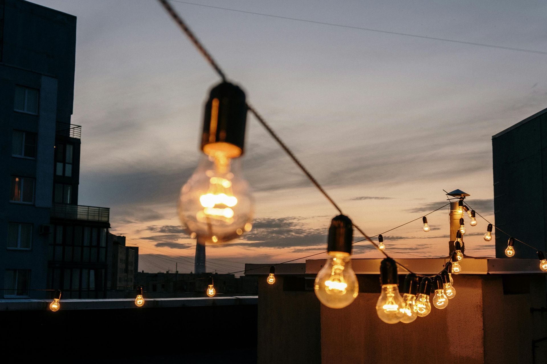 A string of glowing Edison bulbs hanging against a sunset sky, with city buildings visible in the background.