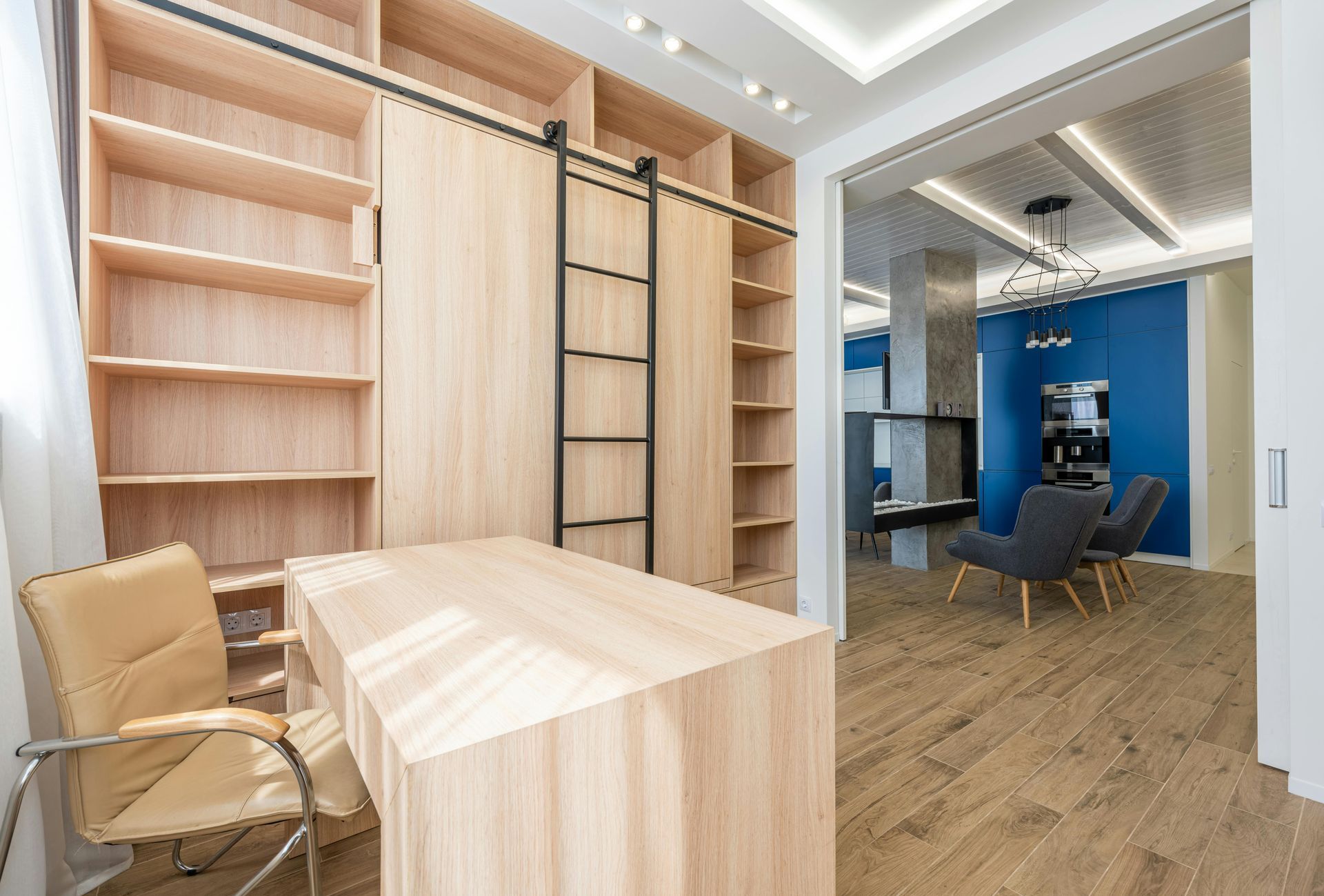 A home office with a light wood desk and bookshelf wall, featuring a sliding ladder, opening into a blue-walled room.