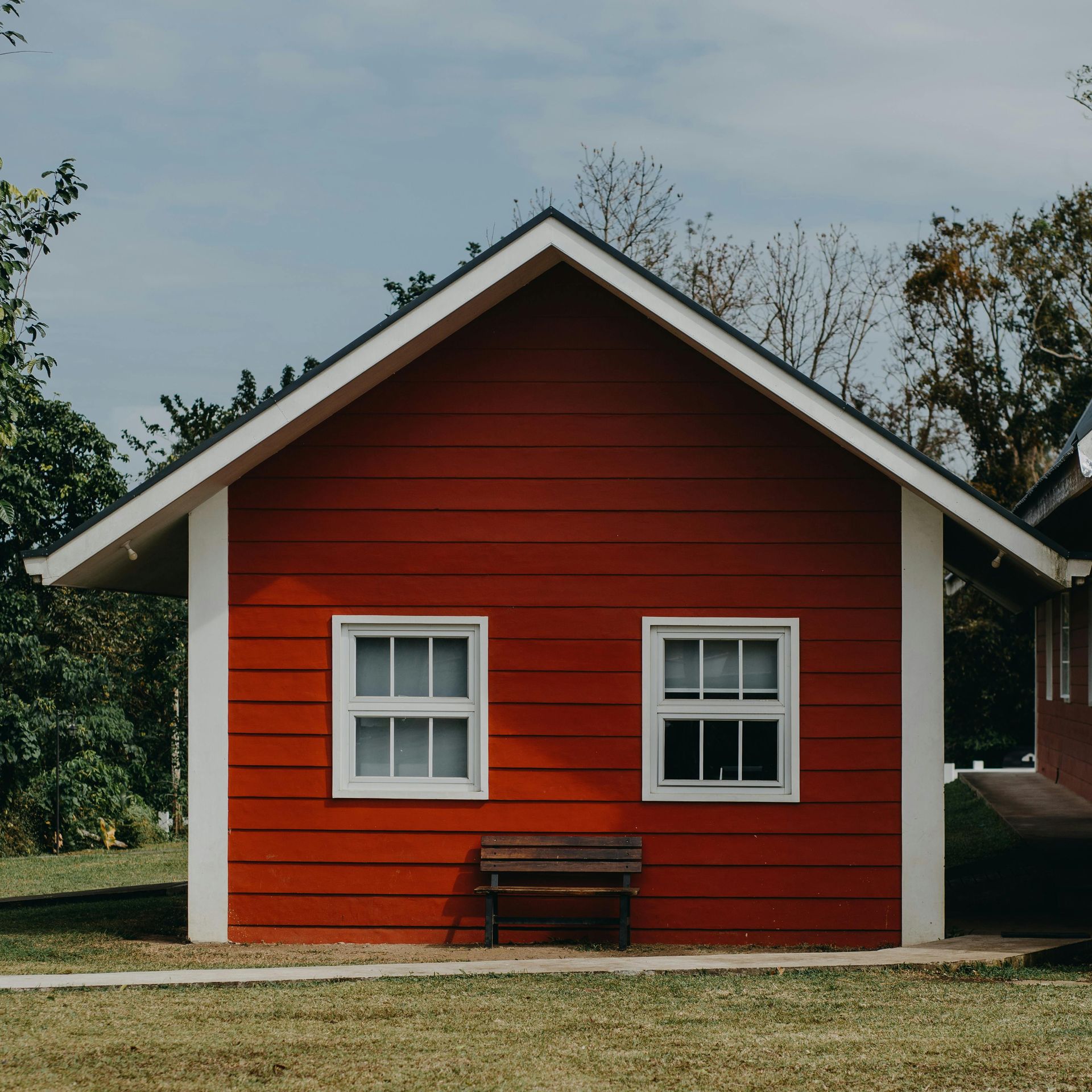 A small, red wooden cabin with a gabled roof, white trim, two windows, and a wooden bench in front, set in a grassy yard.