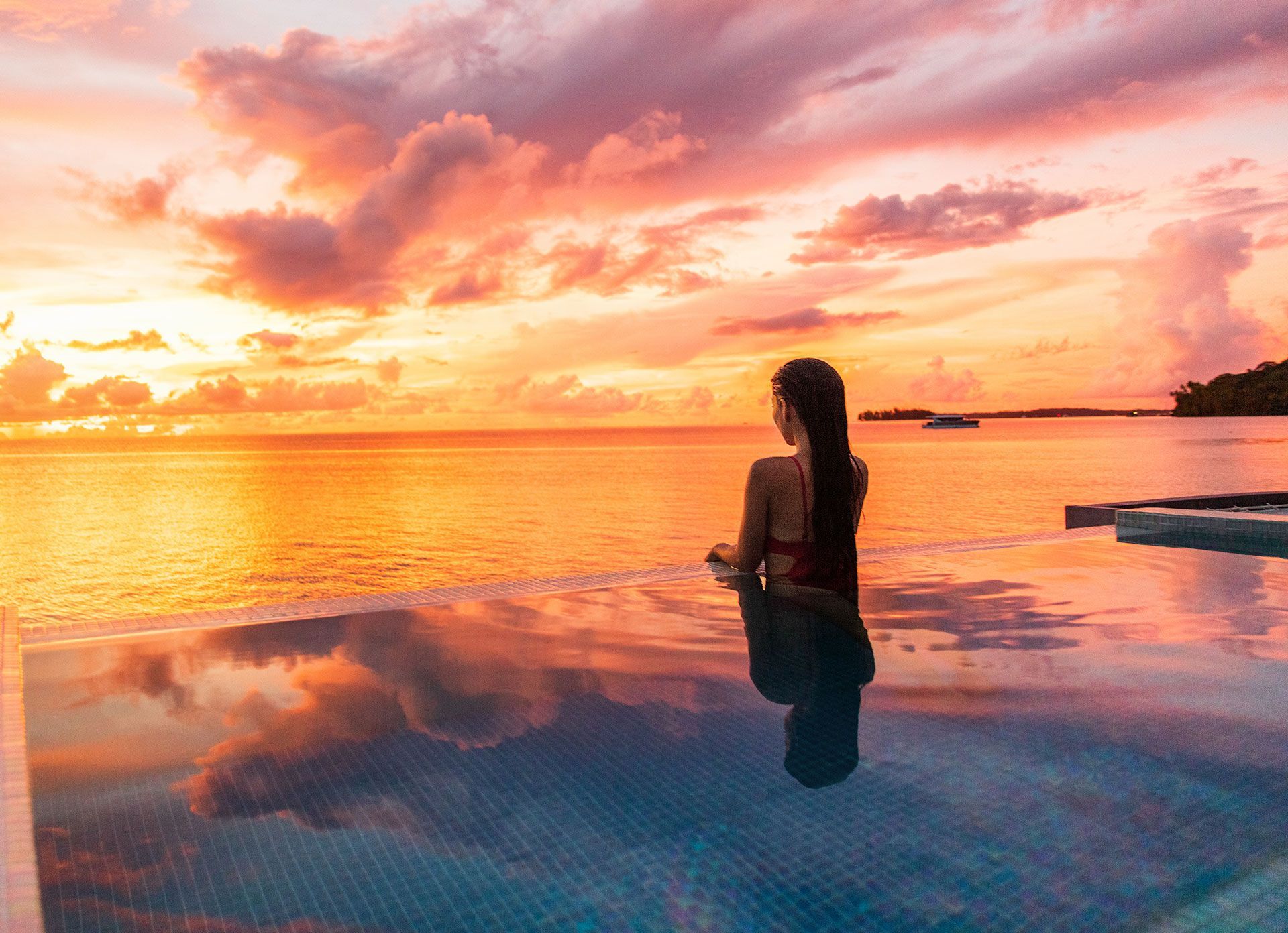 A woman is sitting in an infinity pool overlooking the ocean at sunset.