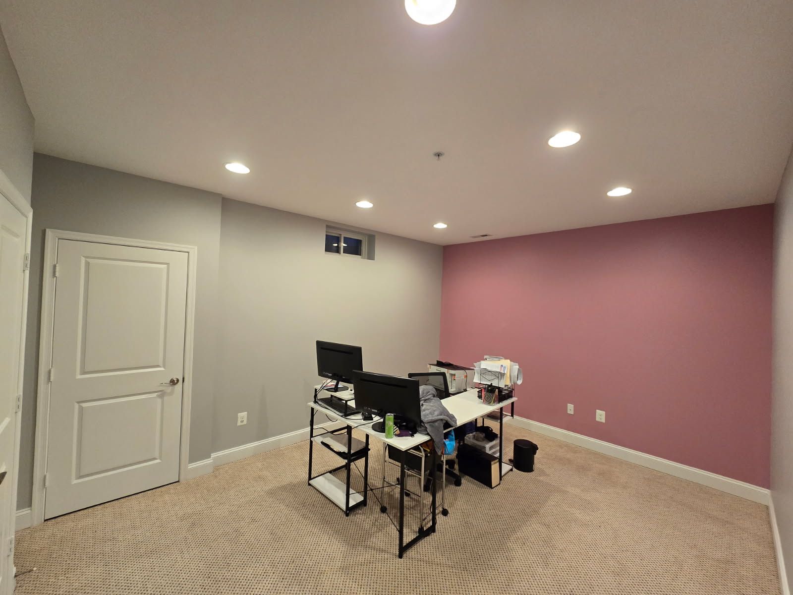 Room with a desk, two computer monitors, gray and pink walls, and a beige patterned carpet.