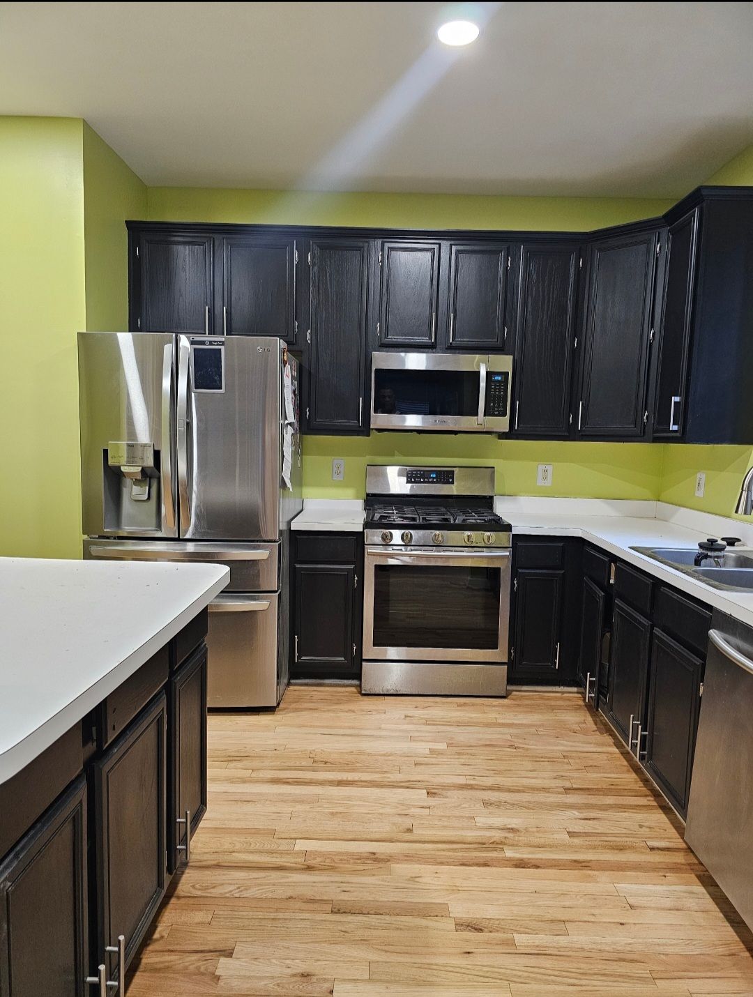 Kitchen with black cabinets, stainless steel appliances, and light wood floors. Green walls and white countertops.