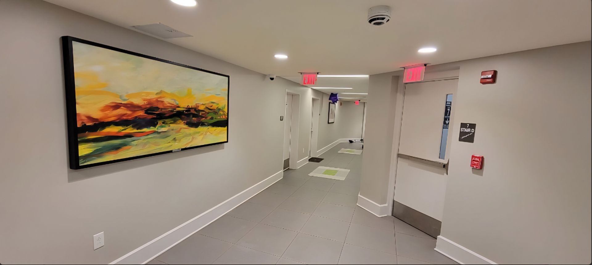 Long hallway with artwork, doors, and exit signs. Grey walls, flooring, and white trim.