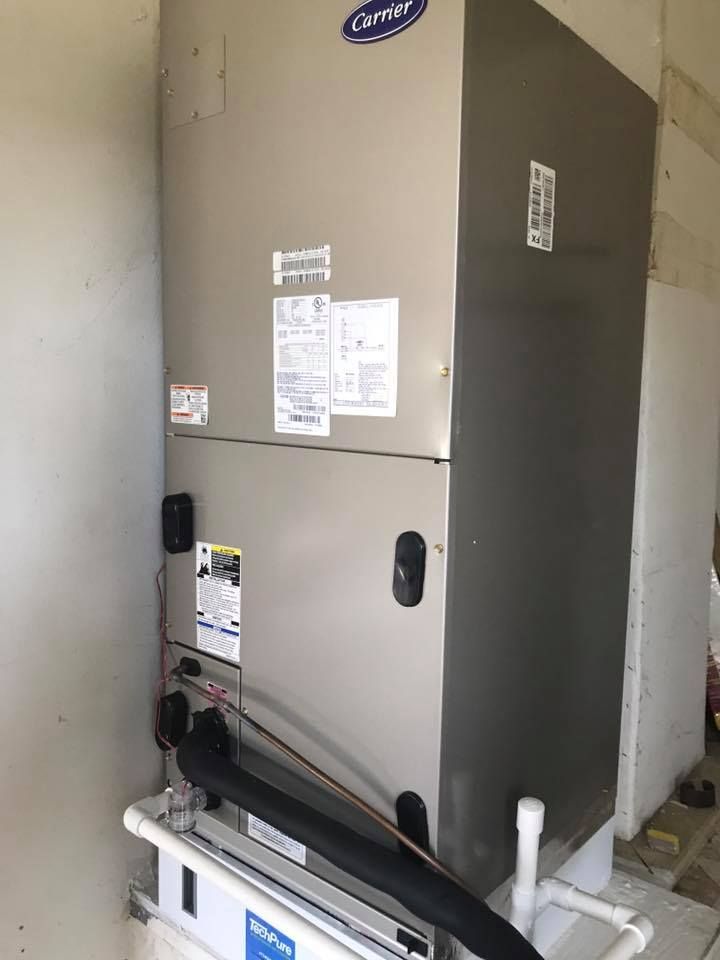 A silver Carrier HVAC unit installed in a utility room. Black insulation and white piping are visible.