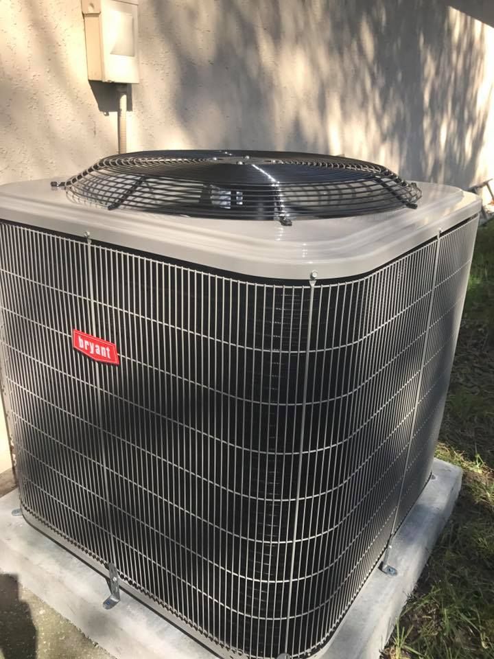 Bryant brand air conditioner unit, gray with black grille, sitting on a concrete pad outside a building.