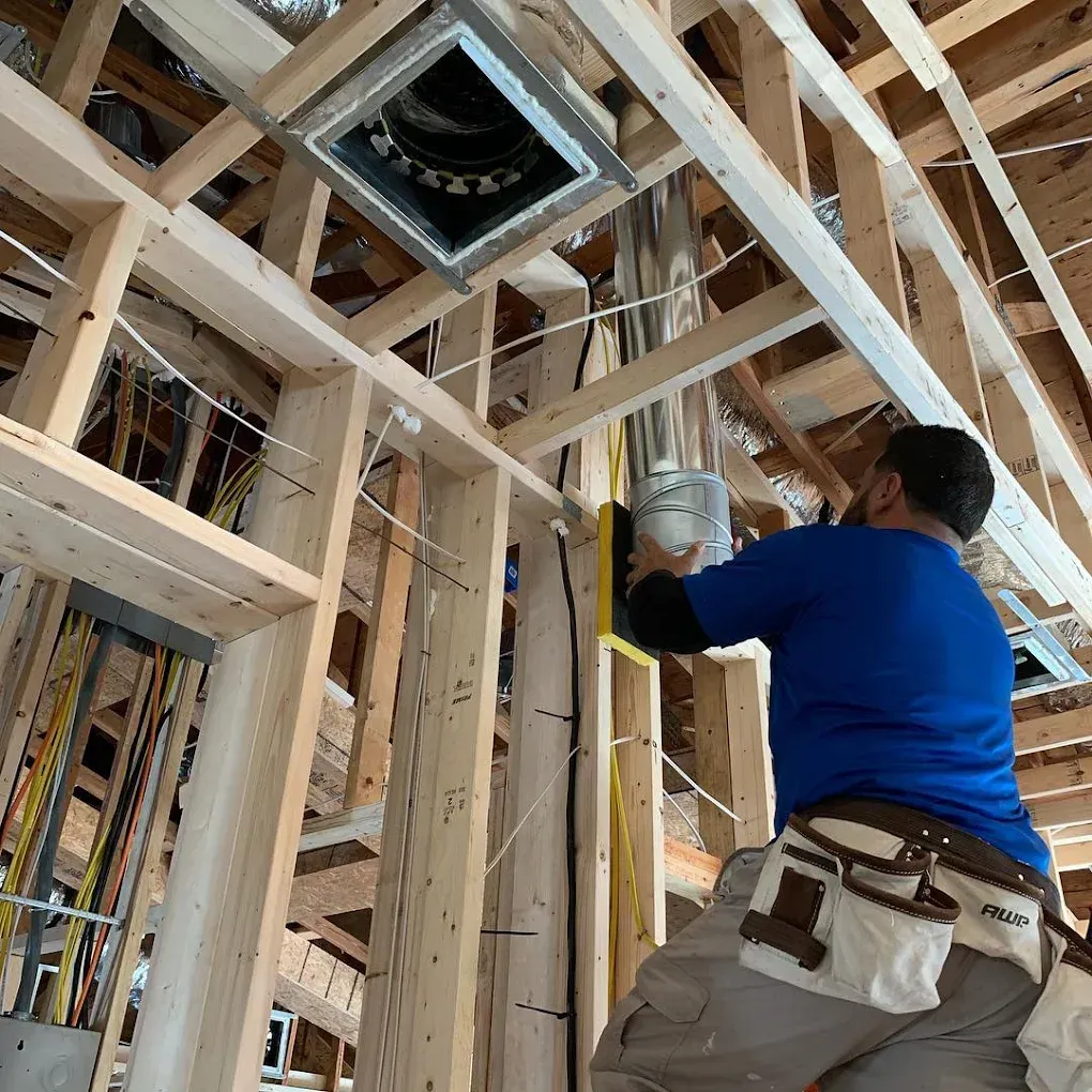 Construction worker installing ductwork in a wood-framed ceiling. He wears a blue shirt, tool belt, and is on a ladder.