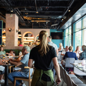 A waitress walks through a busy restaurant with seated patrons. Sunlight streams through large windows.