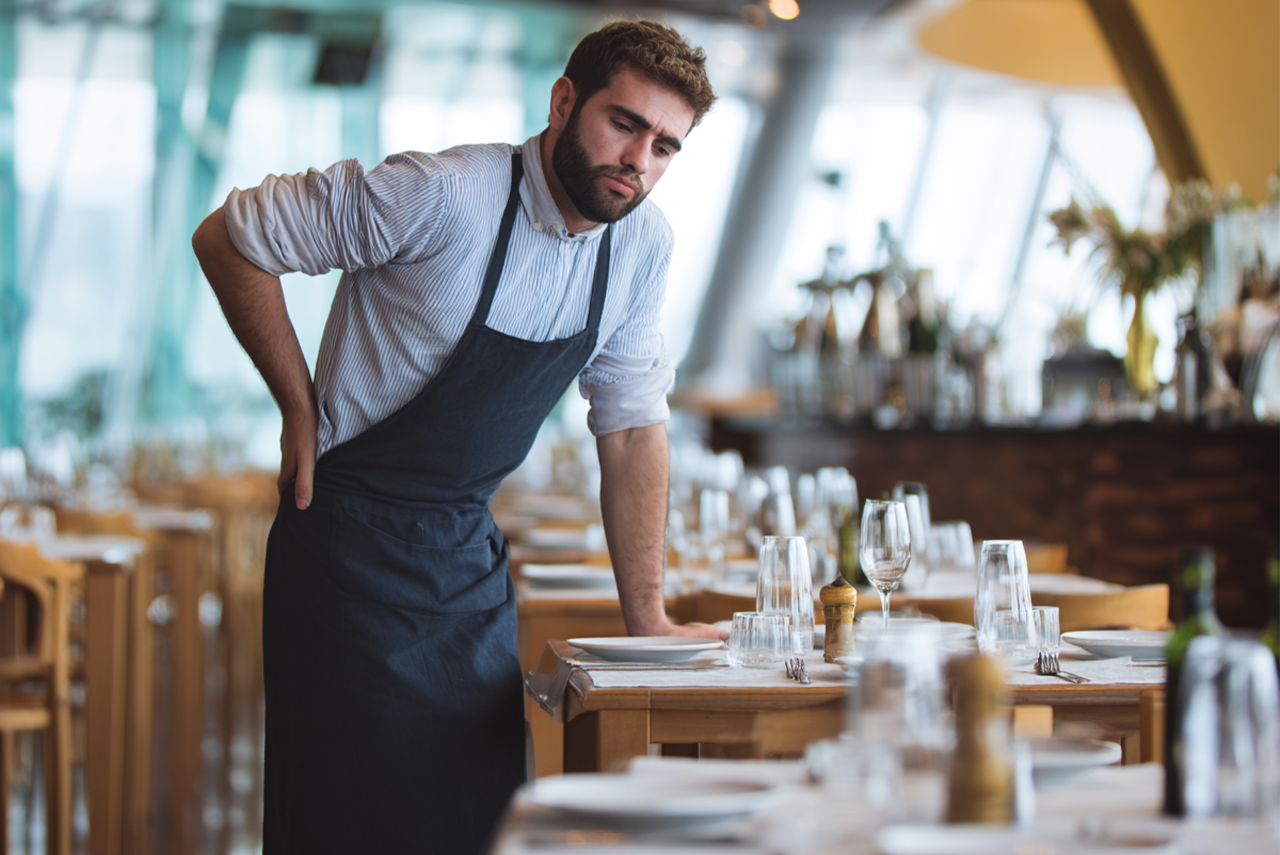 a waiter leans on an empty restaurant table with a look of disappointment on his face