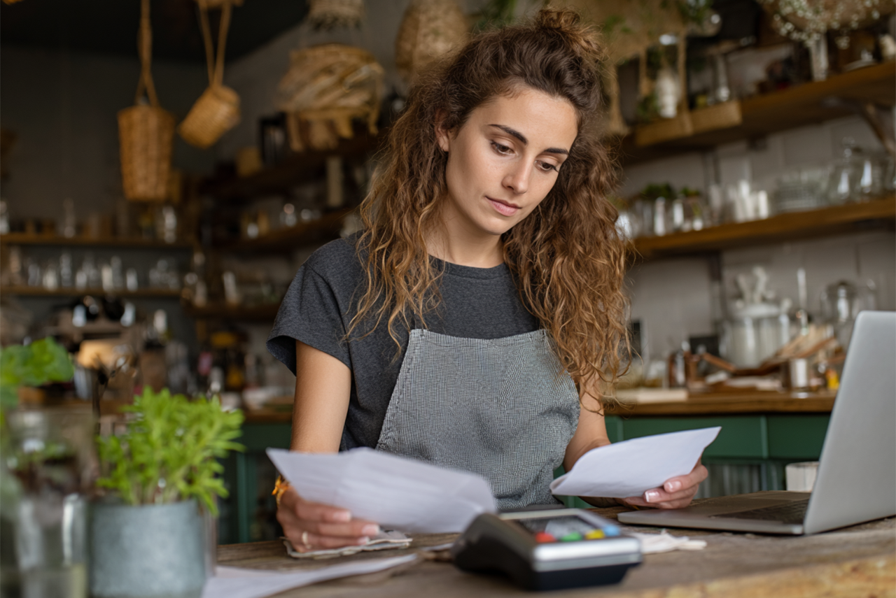 a cafe manager studies two separate bills. Her laptop and card machine are on the table