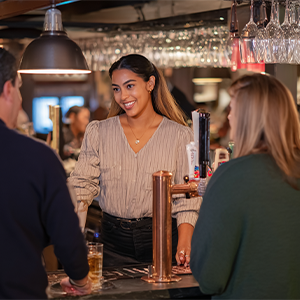 A smiling bartender in a striped shirt serves two customers at a dimly lit bar with glasses hanging overhead.