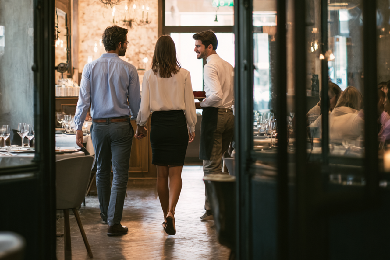 A couple walks towards the exit of a restaurant, chatting with a waiter as they go.