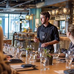 A waiter in a black apron speaks to seated guests around a long wooden table set with wine glasses.