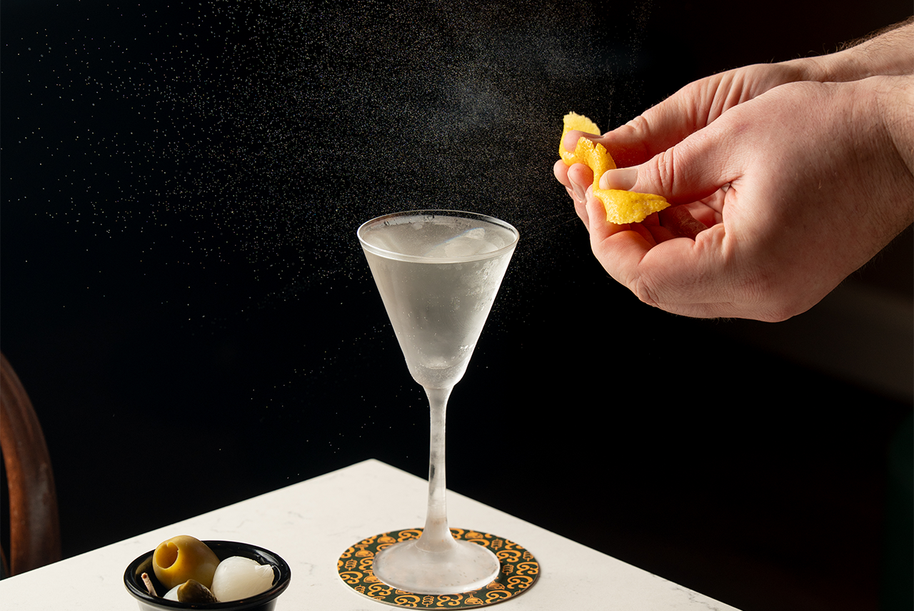 Bartender spritzing citrus oil onto a martini. Martini glass on a coaster, olives on the side.