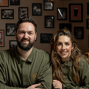 Two people in olive green shirts smile, posing in a bar with framed photos on the wall.