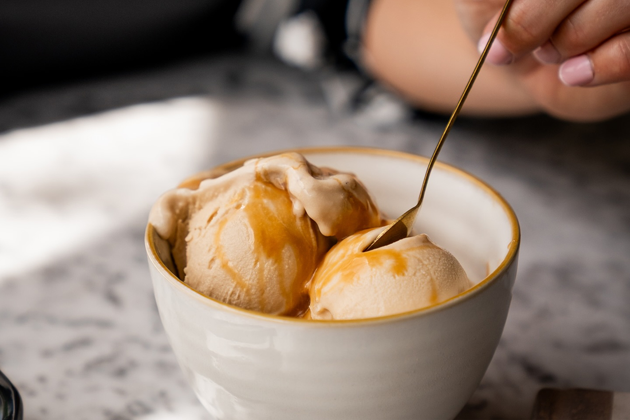 A person’s hand holds a small gold spoon poised over a white bowl filled with scoops of caramel-drizzled ice cream.
