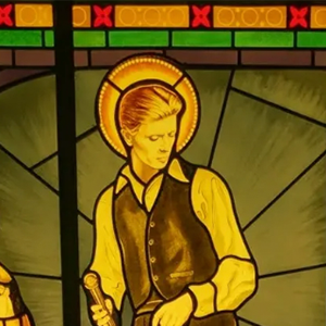 Stained glass of a haloed figure in yellow shirt and dark vest, holding a tool, with geometric background