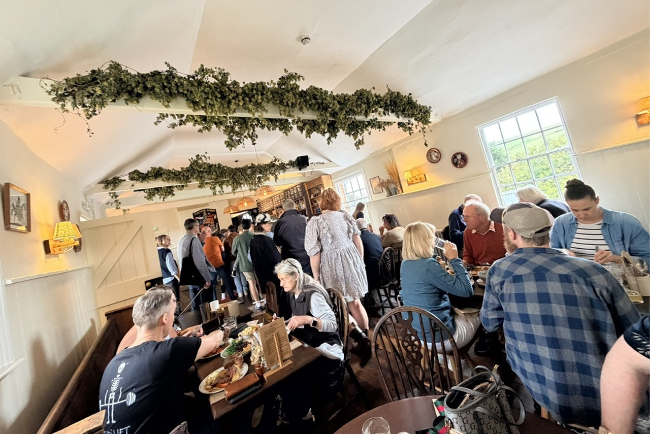 People gathered inside the Trevor Arms, eating at tables. Green vines decorate the ceiling. Bright natural light streams in.