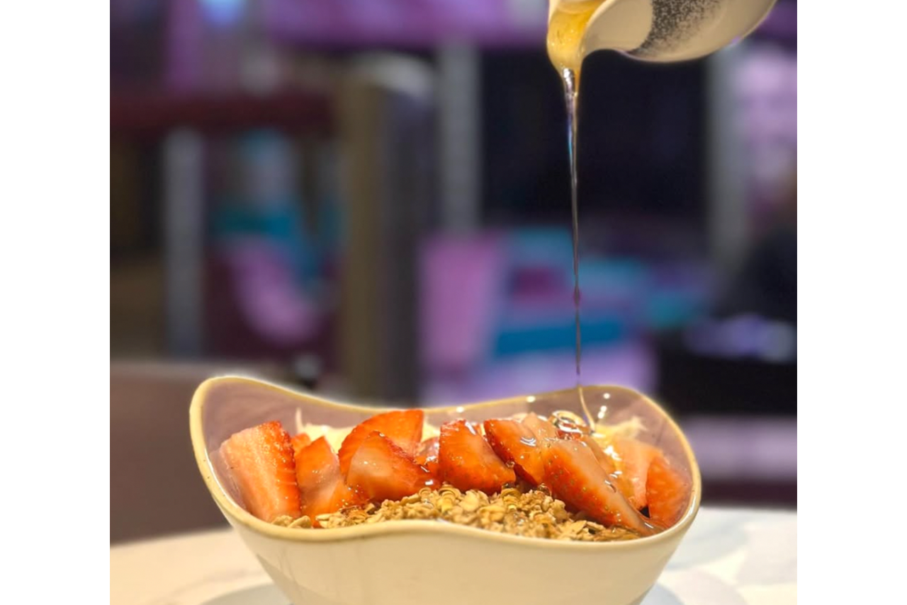 Dessert bowl topped with sliced strawberries and granola as syrup is being poured over it