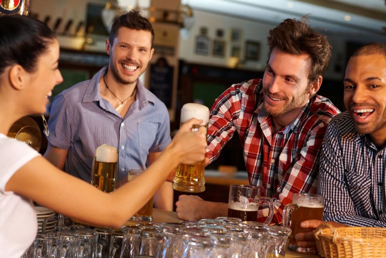 A group of men stand at a bar while a barmaid serves glasses of beer