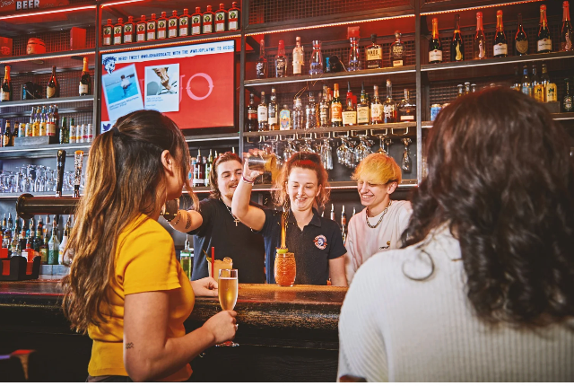 a bartender makes a cocktail while two other barstaff and two guests watch