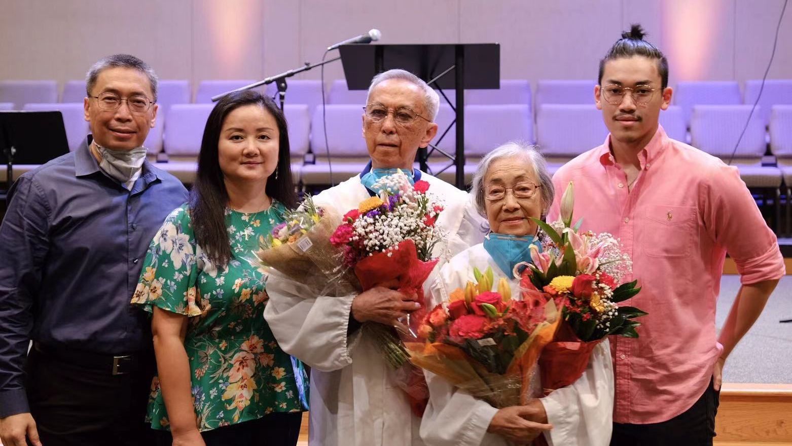 A group of four people standing together on a stage in front of seating, holding bouquets of flowers.