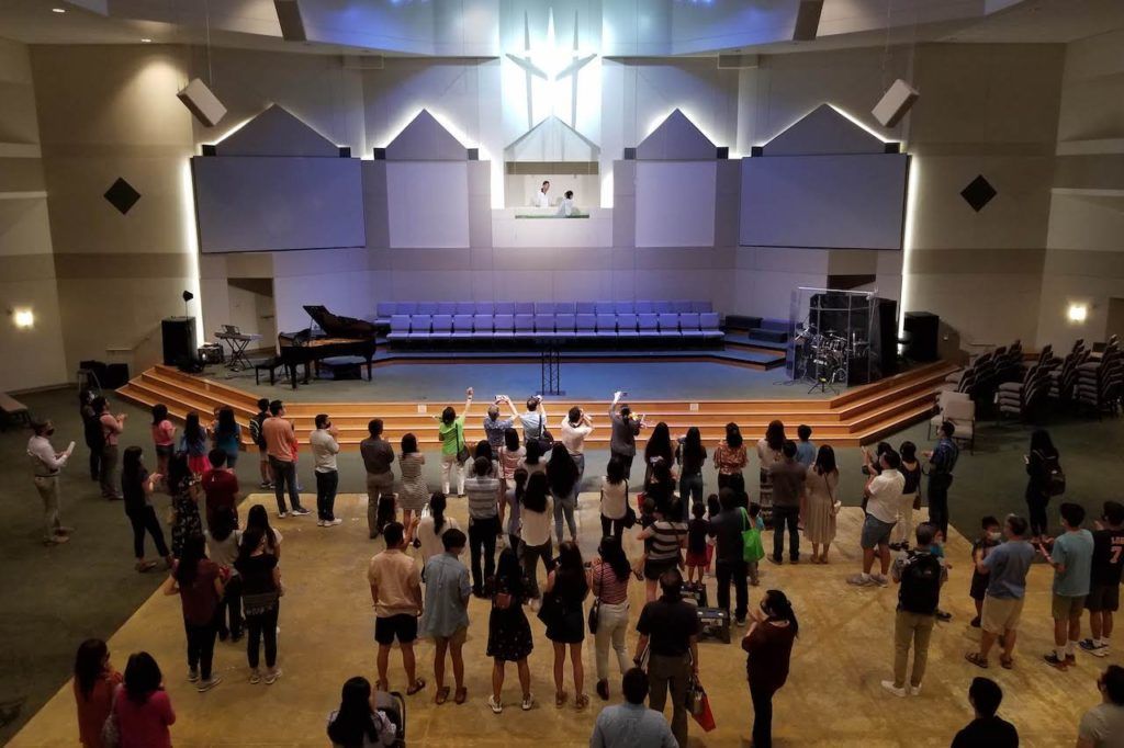 A large group of people stand facing a stage in a brightly lit auditorium during a church service or event.