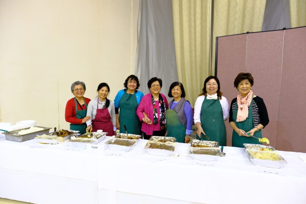 Seven people wearing aprons stand behind a long table filled with serving trays of food in a bright room.