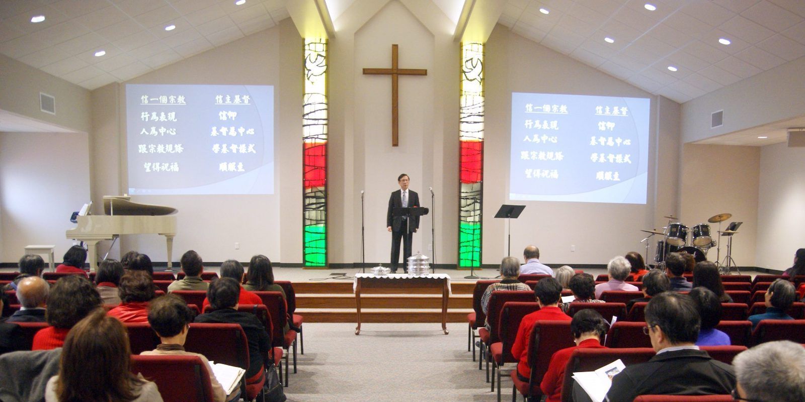 A speaker stands at a podium in a church sanctuary, facing a seated congregation with projection screens on both sides.