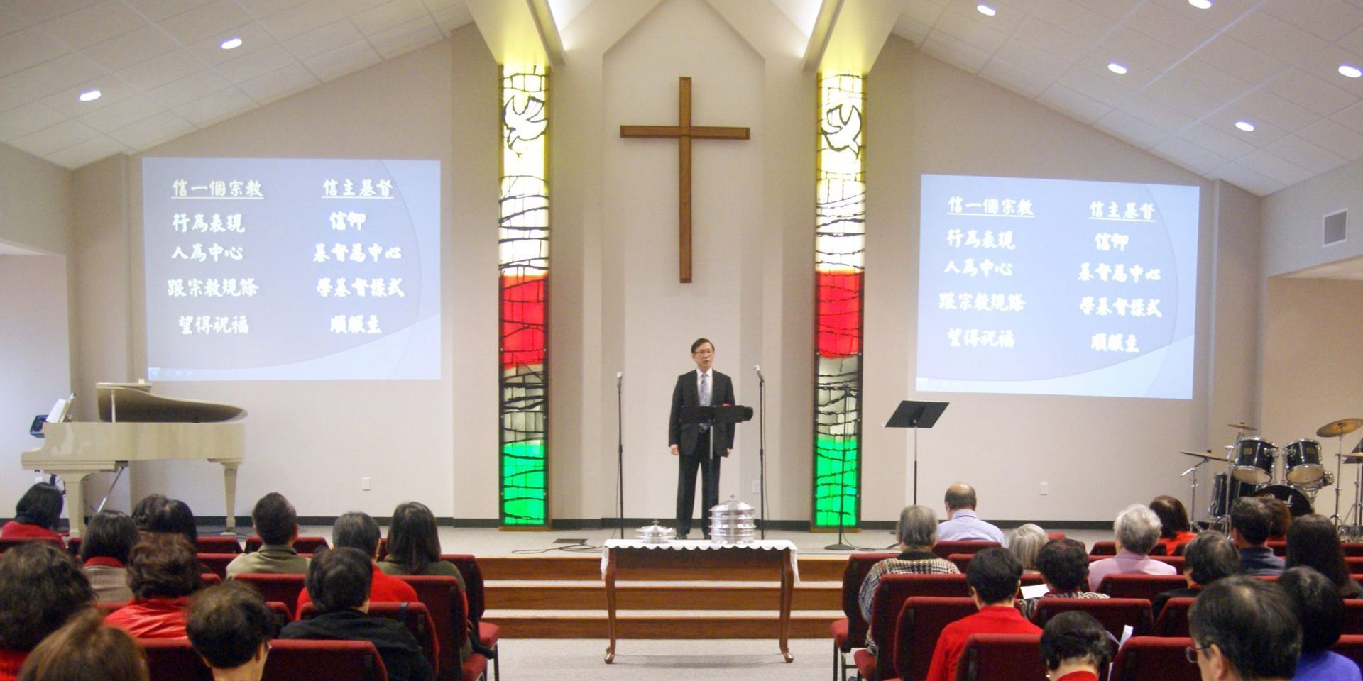 A person in a suit stands at a podium in a church, facing an audience with large screens displaying text on both sides.