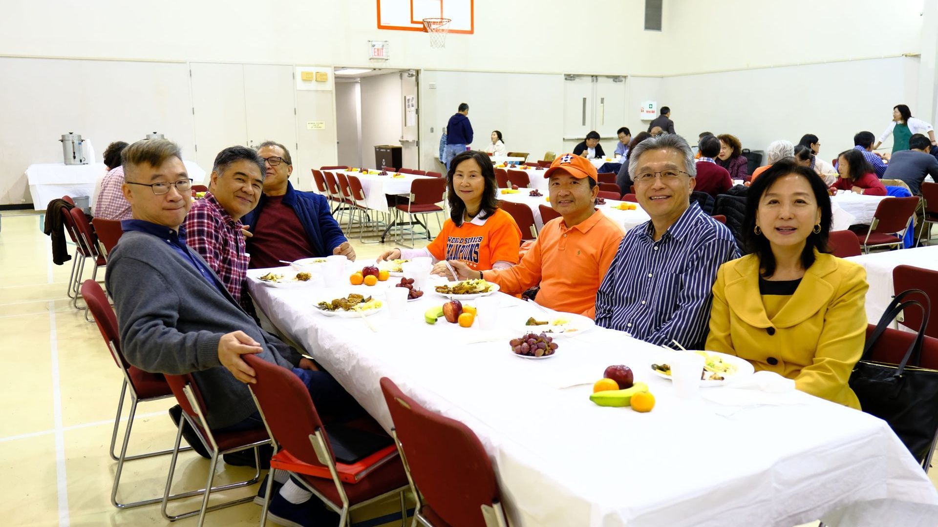 A group of people sit at a long banquet table with white tablecloths and fruit in a brightly lit gymnasium.