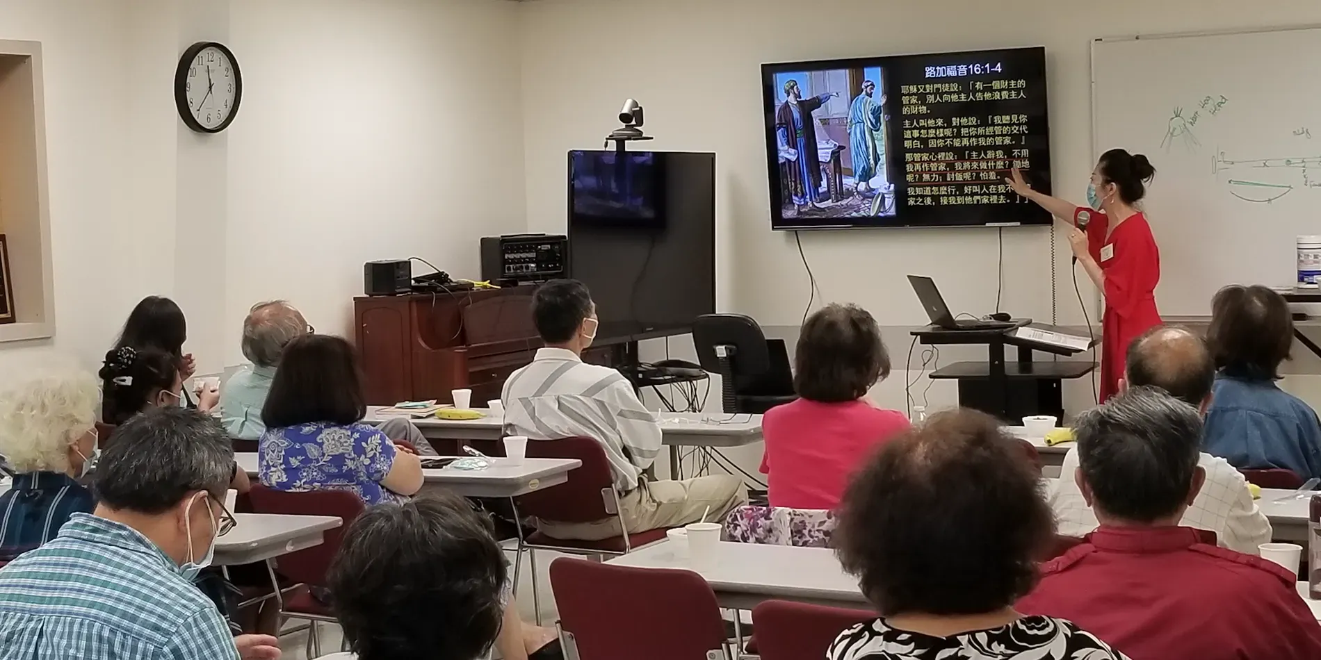 A presenter in a red shirt points to a display screen during a meeting with a group of people in a brightly lit room.