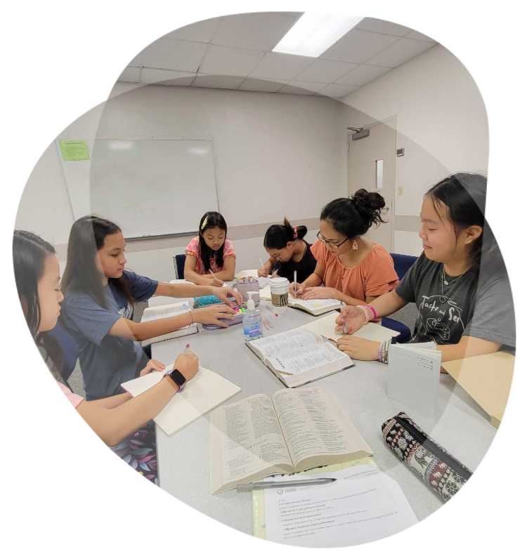 A group of students sits around a table in a classroom, studying and taking notes together from textbooks and notebooks.