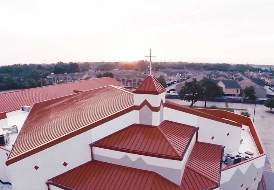 An elevated view of a building with a tiered, reddish-brown metal roof and a steeple topped with a cross at sunset.