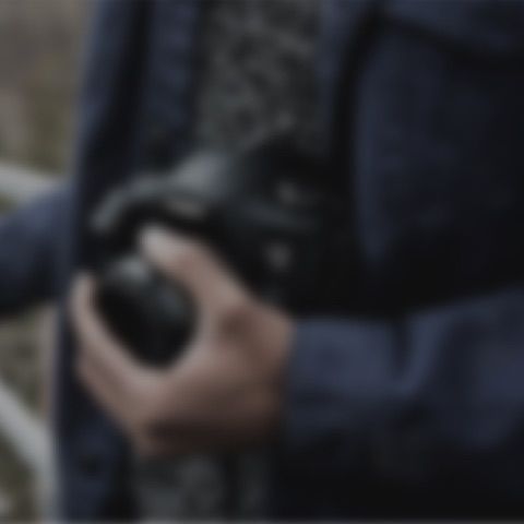 A person in a dark jacket holds a professional camera in their hands, captured in a close-up, out-of-focus shot.