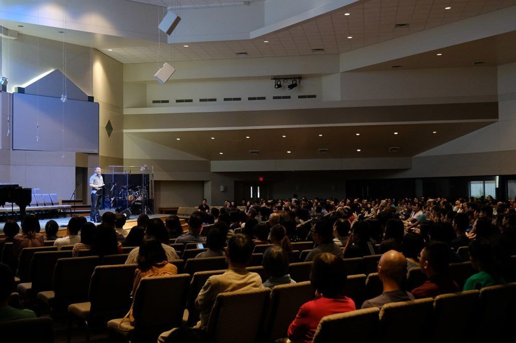 A speaker stands on a stage in a large, dimly lit auditorium addressing a seated audience of many people.
