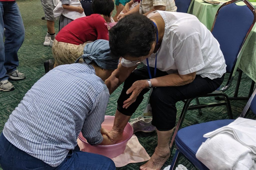 A person in a plaid shirt washes another person's feet in a pink basin while they sit on blue chairs.